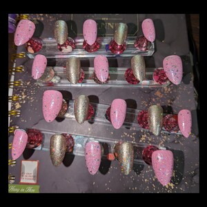 Peut inclure: Un ensemble de vingt faux ongles dispos&eacute;s en quatre rang&eacute;es sur des supports transparents. Les ongles pr&eacute;sentent un m&eacute;lange de rose clair uni avec des paillettes multicolores et une finition dor&eacute;e m&eacute;tallique scintillante, tous en amande.