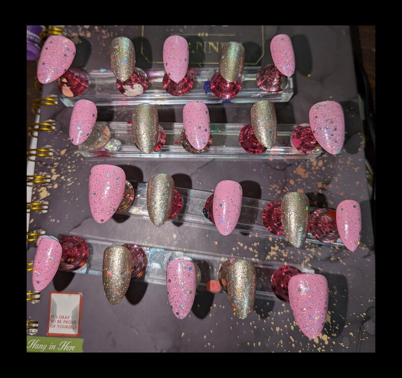 Peut inclure: Un ensemble de vingt faux ongles dispos&eacute;s en quatre rang&eacute;es sur des supports transparents. Les ongles pr&eacute;sentent un m&eacute;lange de rose clair uni avec des paillettes multicolores et une finition dor&eacute;e m&eacute;tallique scintillante, tous en amande.