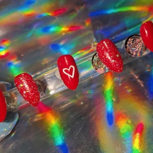 May include: A set of red nails with a glossy finish, including a heart design on one nail and glitter accents on others. The nails are presented on a reflective surface with rainbow light reflections, creating a vibrant aesthetic.