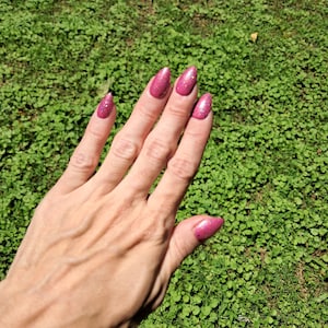 Peut inclure: Une main avec de longs ongles ovales peints avec un vernis rose foncé scintillant. Les ongles ont une finition pailletée. L'arrière-plan est un champ de trèfles verts.