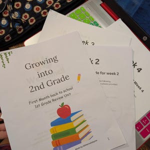 Peut inclure: Une pile de feuilles de travail &eacute;ducatives blanches intitul&eacute;es Growing into 2nd Grade First Month back to school 1st Grade Review Unit. La page de couverture pr&eacute;sente un graphique d'une pomme rouge sur une pile de livres bleu, jaune, vert et orange.