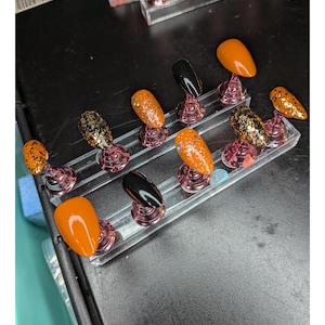 May include: A selection of artificial nails in diverse designs and colours. The nails are presented on clear stands, featuring a variety of styles such as solid orange, black, and glitter-covered options. The arrangement suggests a nail art display or product showcase.