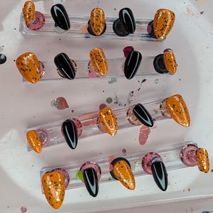 Peut inclure: Une collection de faux ongles de différentes conceptions. Certains ongles sont noirs unis, tandis que d'autres sont orange avec des paillettes dorées et des mouchetures noires. Les ongles sont disposés sur des supports en plastique transparent.