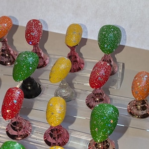 May include: A selection of brightly coloured artificial nails, each coated in glitter, presented on clear acrylic stands with rose gold bases. The nails are in shades of brown, orange, red, yellow, and green.