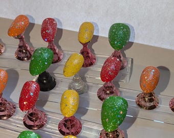 May include: A selection of brightly coloured artificial nails, each coated in glitter, presented on clear acrylic stands with rose gold bases. The nails are in shades of brown, orange, red, yellow, and green.