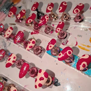 May include: A selection of artificial nails in diverse designs. Some nails are solid red with a white circular design, others are a light pink with red polka dots, and some are covered in red glitter. The nails are displayed on clear plastic stands.