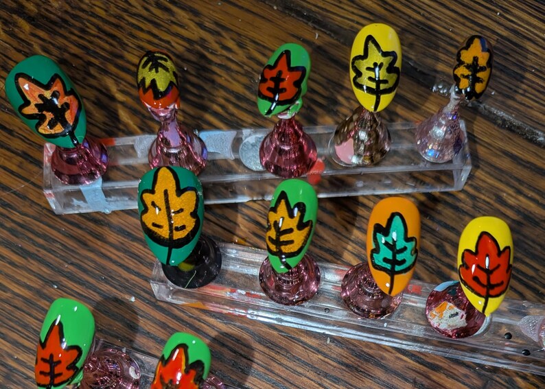 Peut inclure: Un ensemble de dix faux ongles avec des motifs de feuilles d'automne peints &agrave; la main dans des tons vifs de vert, jaune et orange. Chaque ongle est fix&eacute; sur un petit support conique transparent, mettant en valeur des contours noirs d&eacute;taill&eacute;s.