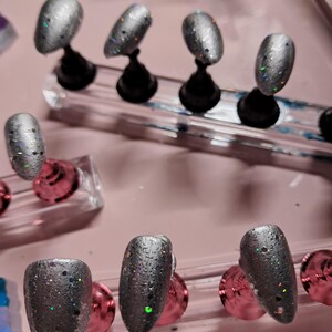 Peut inclure: Des ongles à pression argentés à paillettes sur des supports noirs. Les ongles ont une finition texturée et sont ornés de paillettes irisées. Les ongles sont disposés sur une surface rose, présentant un ensemble d'ongles artificiels.