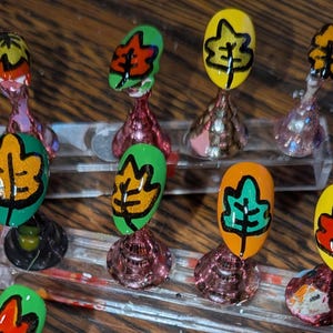 Peut inclure: Un ensemble de dix faux ongles mont&eacute;s sur des supports en acrylique transparent. Chaque ongle pr&eacute;sente un motif de feuille peint &agrave; la main dans des couleurs vives comme le vert, le jaune, l'orange et le rouge, soulign&eacute; de noir.
