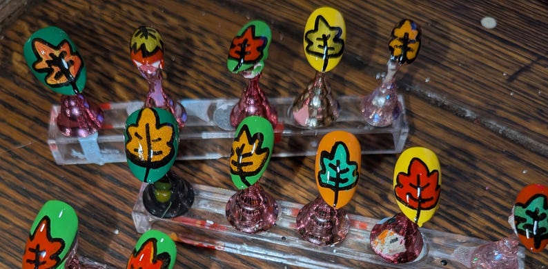 Peut inclure: Un ensemble de dix faux ongles mont&eacute;s sur des supports en acrylique transparent. Chaque ongle pr&eacute;sente un motif de feuille peint &agrave; la main dans des couleurs vives comme le vert, le jaune, l'orange et le rouge, soulign&eacute; de noir.