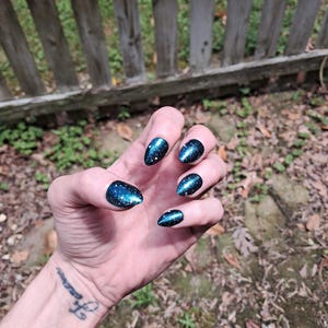 Peut inclure: Une main avec de longs ongles en amande peints en bleu foncé avec un effet pailleté rappelant une galaxie. Les ongles ont une finition brillante. L'arrière-plan comprend une clôture en bois et du feuillage.