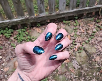 May include: A hand with long, almond-shaped nails painted in a dark blue color with a glittery, galaxy-like effect. The nails have a glossy finish, reflecting light. The background includes a wooden fence and foliage.