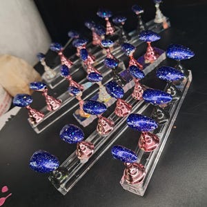 Peut inclure: Collection de faux ongles bleu foncé pailletés, présentés sur des présentoirs en acrylique transparent. Les présentoirs ont des accents or rose et argent. Idéal pour un salon de manucure ou une présentation de produits.
