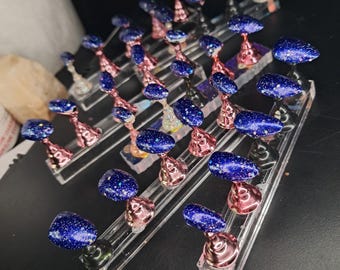 May include: A collection of artificial nails with a dark blue glitter finish. The nails are displayed on clear acrylic stands with rose gold and silver accents. The arrangement suggests a nail art display or product showcase.