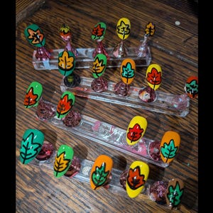 Peut inclure: Un ensemble de faux ongles d&eacute;cor&eacute;s de motifs de feuilles d'automne peints &agrave; la main dans des tons vifs de vert, jaune et orange. Chaque ongle pr&eacute;sente un contour noir marqu&eacute;, dispos&eacute; sur des supports en acrylique transparent pour la pr&eacute;sentation.
