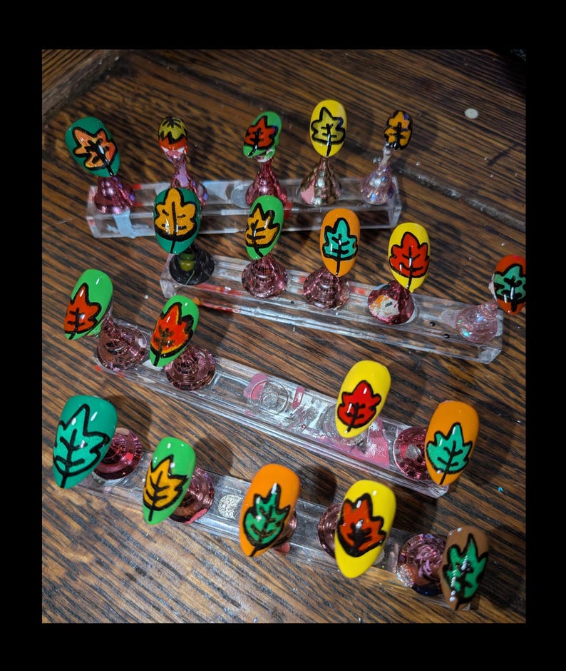Peut inclure: Un ensemble de faux ongles d&eacute;cor&eacute;s de motifs de feuilles d'automne peints &agrave; la main dans des tons vifs de vert, jaune et orange. Chaque ongle pr&eacute;sente un contour noir marqu&eacute;, dispos&eacute; sur des supports en acrylique transparent pour la pr&eacute;sentation.