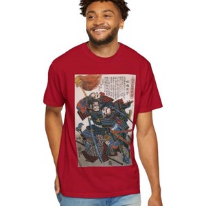 以下が含まれることがあります： 赤色のTシャツ。日本の浮世絵風のプリントで、戦闘中の侍が描かれています。プリントには、鎧を着た詳細な人物、刀、日本語のテキストが含まれています。Tシャツは無地の赤色です。