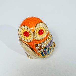 May include: A decorative owl-shaped trinket box featuring a bright orange head, cream-colored body, and two large eyes with white petals and red crystal centers. The wings are embellished with clear rhinestones and blue and purple enamel accents.