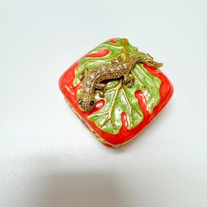 May include: A decorative trinket box with a red and green leaf design. A gold-colored lizard with crystal accents rests on top. The box has a square shape and a gold-colored trim.