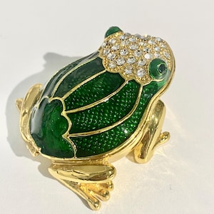 May include: A gold-toned frog-shaped brooch with a vibrant green enamel body and sparkling crystal accents. The frog's eyes are green, and the brooch has a detailed, textured design. The brooch is a decorative accessory.