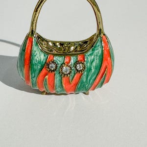 May include: A miniature handbag-shaped trinket box. The box is green with red stripes and three clear, round gemstones. The handle and trim are gold-colored. The trinket box has small gold feet.