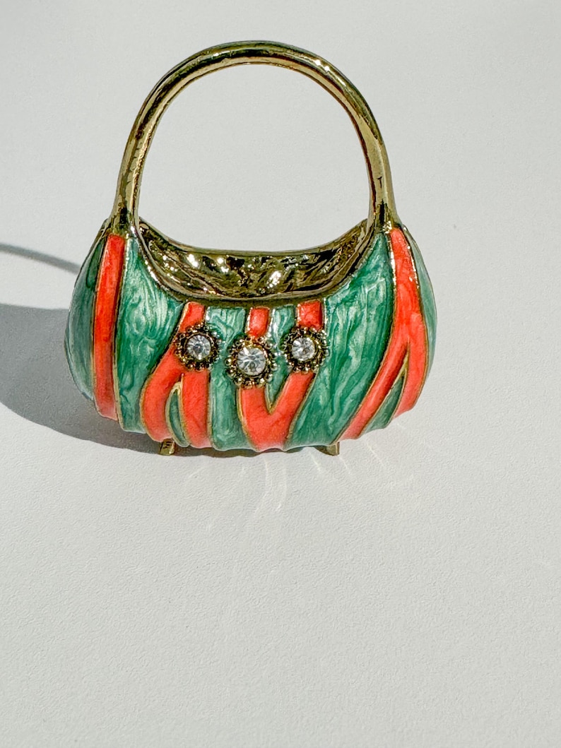 May include: A miniature handbag-shaped trinket box. The box is green with red stripes and three clear, round gemstones. The handle and trim are gold-colored. The trinket box has small gold feet.
