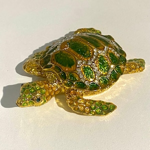May include: A decorative turtle figurine with a gold-colored body and green shell. The shell is adorned with sparkling clear crystals. The turtle's head and flippers are detailed with green and gold accents.