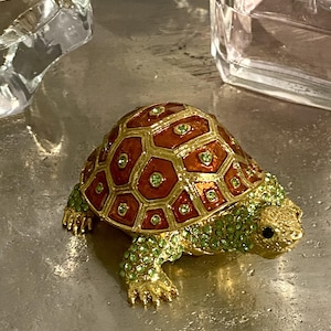 May include: A decorative turtle figurine with a red and gold shell, green jeweled accents, and gold-colored feet and head. The turtle is a small, detailed ornament.