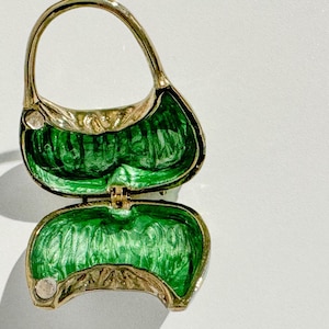 May include: A gold-toned, open purse-shaped trinket box with a handle. The interior is a vibrant green, and the edges are detailed with a leaf-like pattern. The box is hinged and appears to be made of metal.