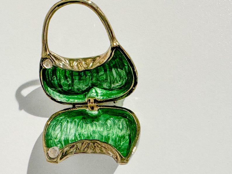 May include: A gold-toned, open purse-shaped trinket box with a handle. The interior is a vibrant green, and the edges are detailed with a leaf-like pattern. The box is hinged and appears to be made of metal.