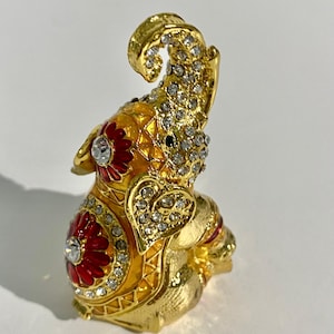 May include: A gold-colored elephant figurine with intricate detailing. The elephant is adorned with red and white accents and sparkling rhinestones. The trunk is raised, and the elephant is in a sitting position. The figurine has a decorative, ornate design.