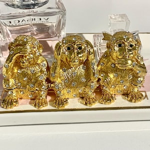 May include: Three gold-colored monkey figurines in the 'see no evil, hear no evil, speak no evil' pose. Each monkey is adorned with small, clear crystals. The figurines are displayed on a white and pink tray, with perfume bottles in the background.