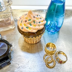 May include: A decorative cupcake-shaped trinket box with a gold base and a brown top, topped with sprinkles. Beside it are gold rings, a blue perfume bottle, a watch, and a perfume bottle with a crystal top, all on a silver tray.