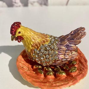 May include: A decorative hen figurine with a red comb and yellow and purple feathers, adorned with sparkling rhinestones. The hen sits on a nest with golden eggs, all set on an orange base. A charming, detailed collectible.