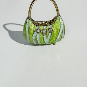 May include: A miniature handbag with a gold handle and trim. The body of the bag is green and light blue striped, with three clear, round embellishments. The bag has small gold feet.