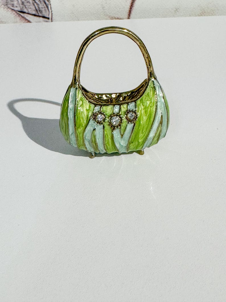 May include: A miniature handbag with a gold handle and trim. The body of the bag is green and light blue striped, with three clear, round embellishments. The bag has small gold feet.