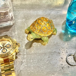 May include: A decorative turtle figurine with a gold shell and green accents. The turtle is on a silver tray with a gold wristwatch, perfume bottle, and a blue bottle.