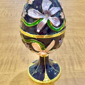 May include: A decorative, navy blue egg-shaped trinket box with a gold base and accents. The box is adorned with lavender and peach-colored floral designs, accented with green and gold trim. The egg sits on a matching pedestal.