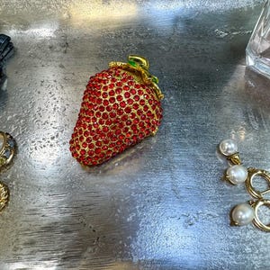 May include: A gold-toned strawberry-shaped brooch encrusted with red rhinestones. The brooch has a gold stem and green leaves. The brooch is on a silver surface, with other jewelry pieces visible in the background.