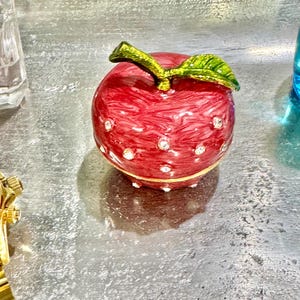 May include: A red apple-shaped trinket box with a green leaf and stem. The box is embellished with small, sparkling rhinestones and a gold-colored band. The decorative box is a charming accent piece.