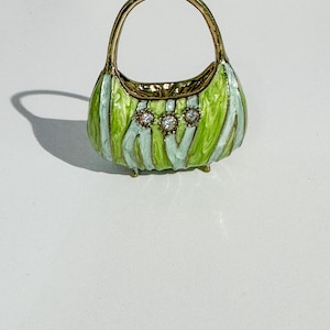 May include: A miniature decorative handbag with a gold handle and trim. The body of the bag is green with light blue stripes and three clear, round gemstones. The bag is approximately 2 inches tall.