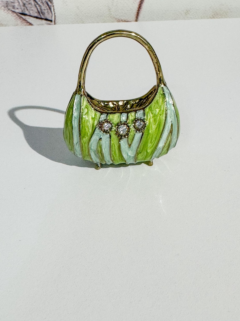 May include: A miniature decorative handbag with a gold handle and trim. The body of the bag is green with light blue stripes and three clear, round gemstones. The bag is approximately 2 inches tall.