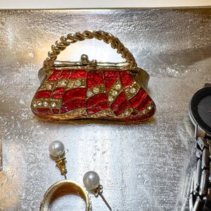 May include: A small, decorative purse with a red and gold geometric design, embellished with rhinestones. The purse has a twisted gold handle. Also pictured are gold earrings with pearls, a gold ring, and a watch.