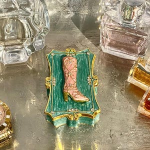 May include: A decorative trinket box featuring a pink and gold cowboy boot design. The boot is set against a green background within a gold-trimmed frame. The box is embellished with rhinestones and sits on a silver surface, surrounded by perfume bottles.