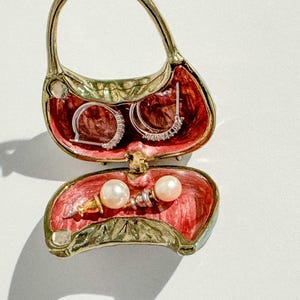 May include: A decorative handbag-shaped jewelry box, open to reveal earrings and pearls. The interior is red, and the exterior is gold and green. The earrings are silver with clear stones. The pearls are white.