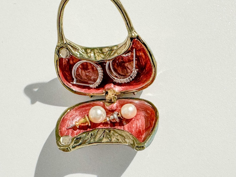 May include: A decorative handbag-shaped jewelry box, open to reveal earrings and pearls. The interior is red, and the exterior is gold and green. The earrings are silver with clear stones. The pearls are white.