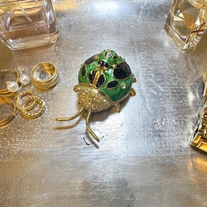 May include: A decorative ladybug trinket box with green enamel, black spots, and crystal accents. The ladybug has gold-colored legs and antennae. Other jewelry and perfume bottles are also visible.