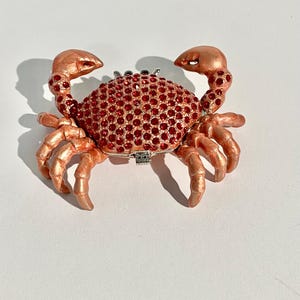 May include: A decorative crab figurine with a metallic bronze finish. The crab's body is covered in small, red rhinestones. The claws and legs are bronze. The crab is a decorative item.