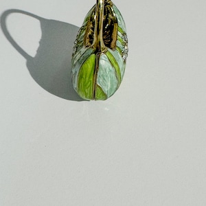 May include: A decorative swan-shaped trinket box. The box is painted in shades of green and white, with gold-colored accents and small, sparkling embellishments. The swan's neck forms a handle.