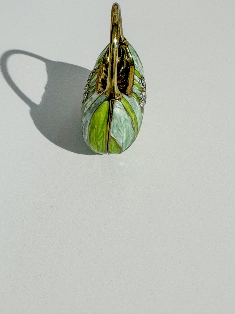 May include: A decorative swan-shaped trinket box. The box is painted in shades of green and white, with gold-colored accents and small, sparkling embellishments. The swan's neck forms a handle.